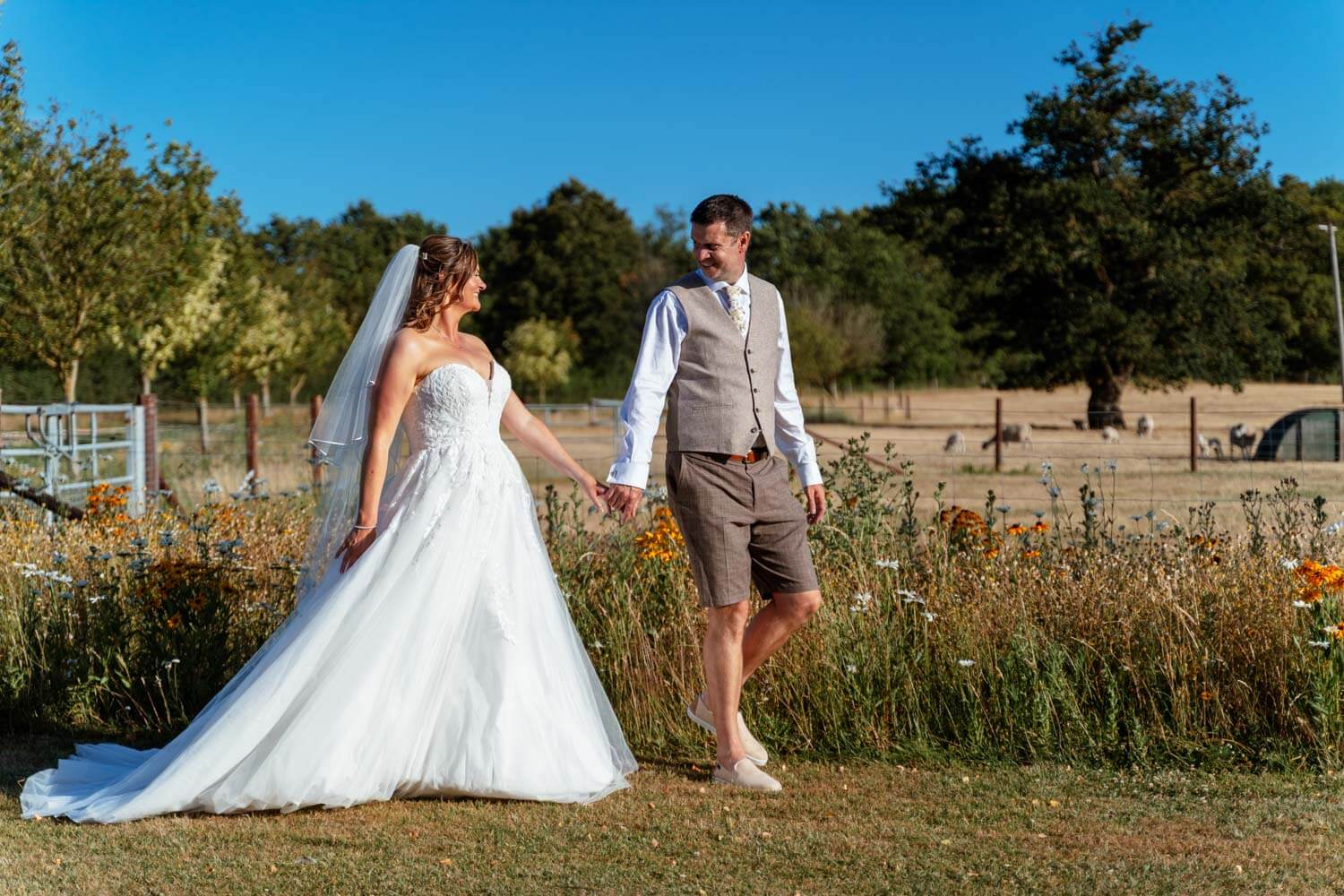 The bride and groom walk hand in hand through a wildflower meadow with sheep grazing behind them.