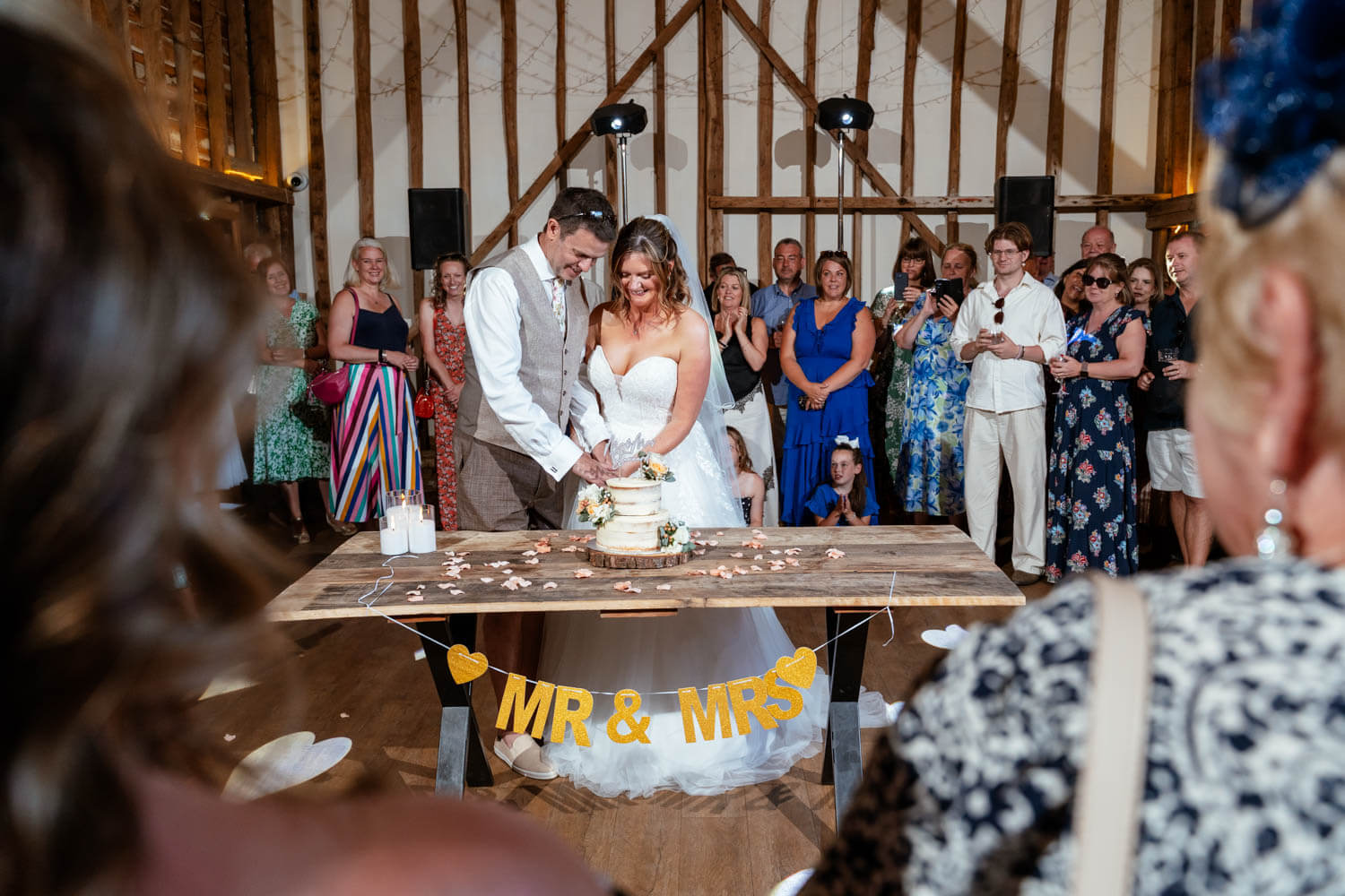 The bride and groom cut their wedding cake inside Stokes Farm Barn, guests gathered around.