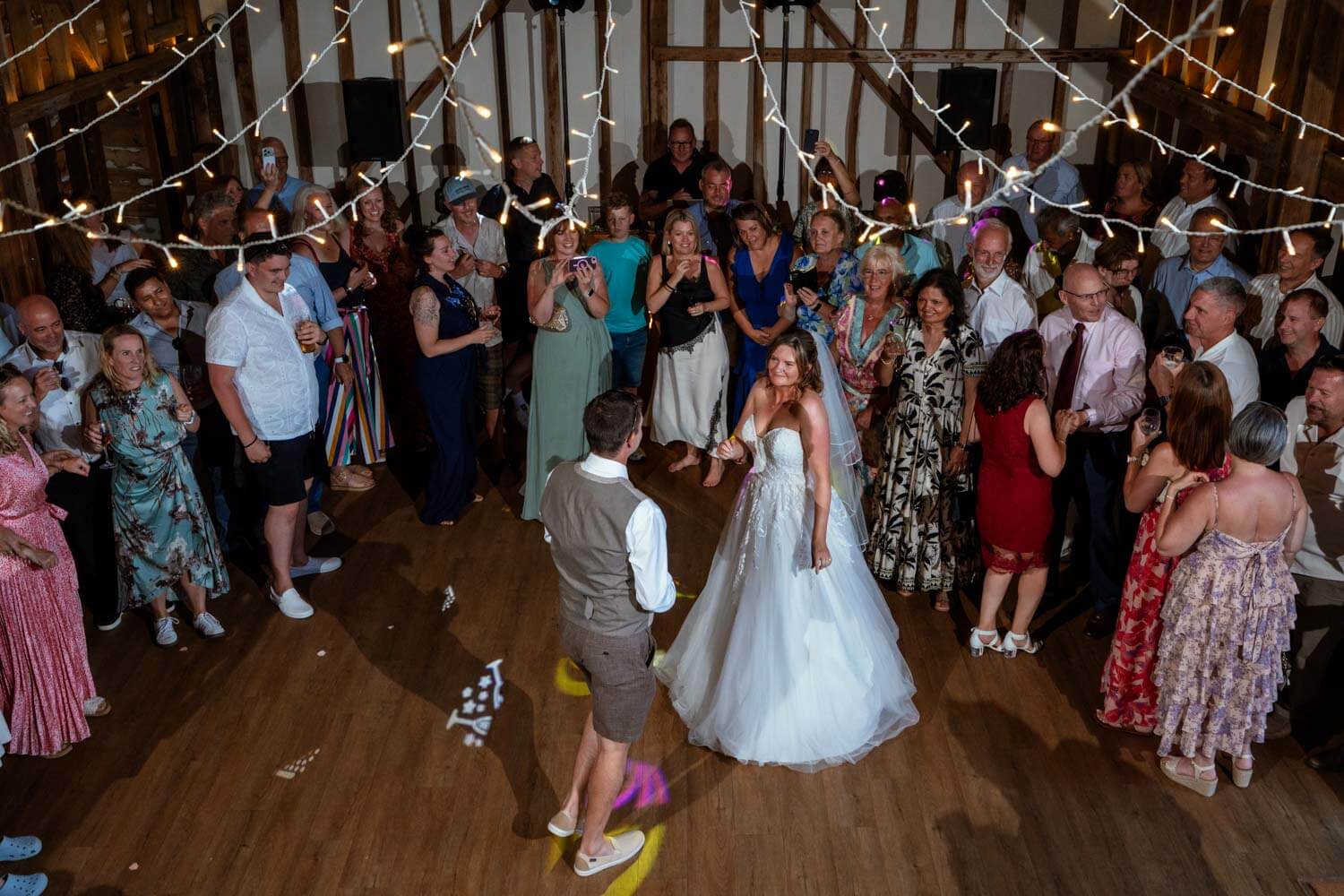 The bride and groom's first dance, surrounded by guests, under fairy lights strung across the barn beams.