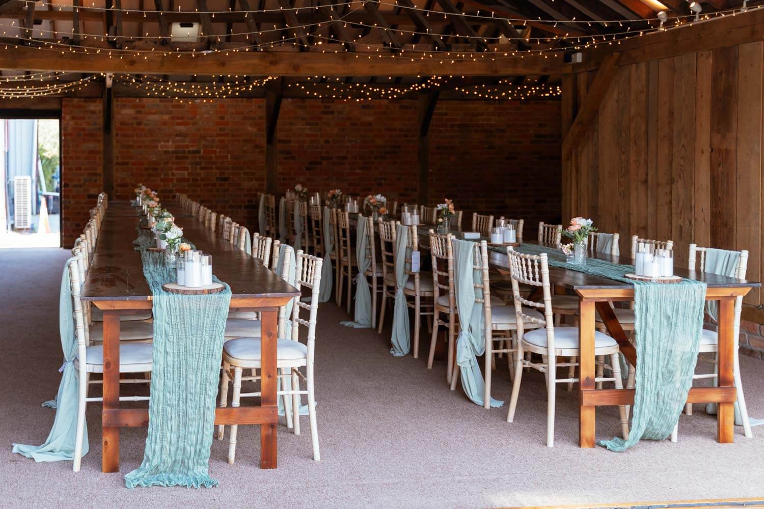 Long reception tables are dressed with sage runners inside the barn, fairy lights strung across the beams.