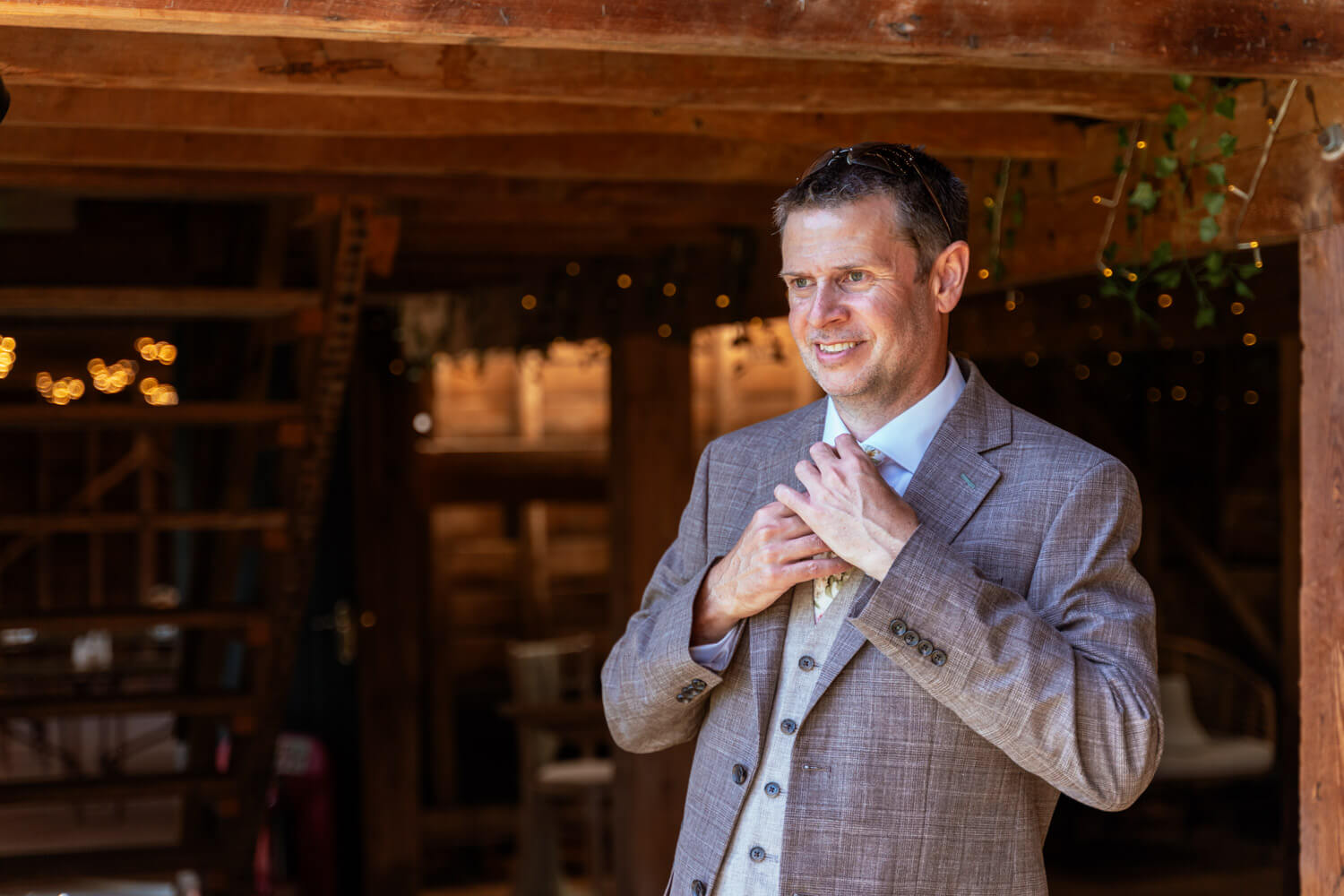 Groom adjusting his tie under the timber beams of the barn.